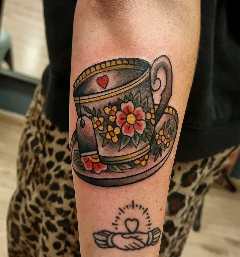 Traditionele tattoo van een theekop met bloemen en een Claddagh symbool eronder.