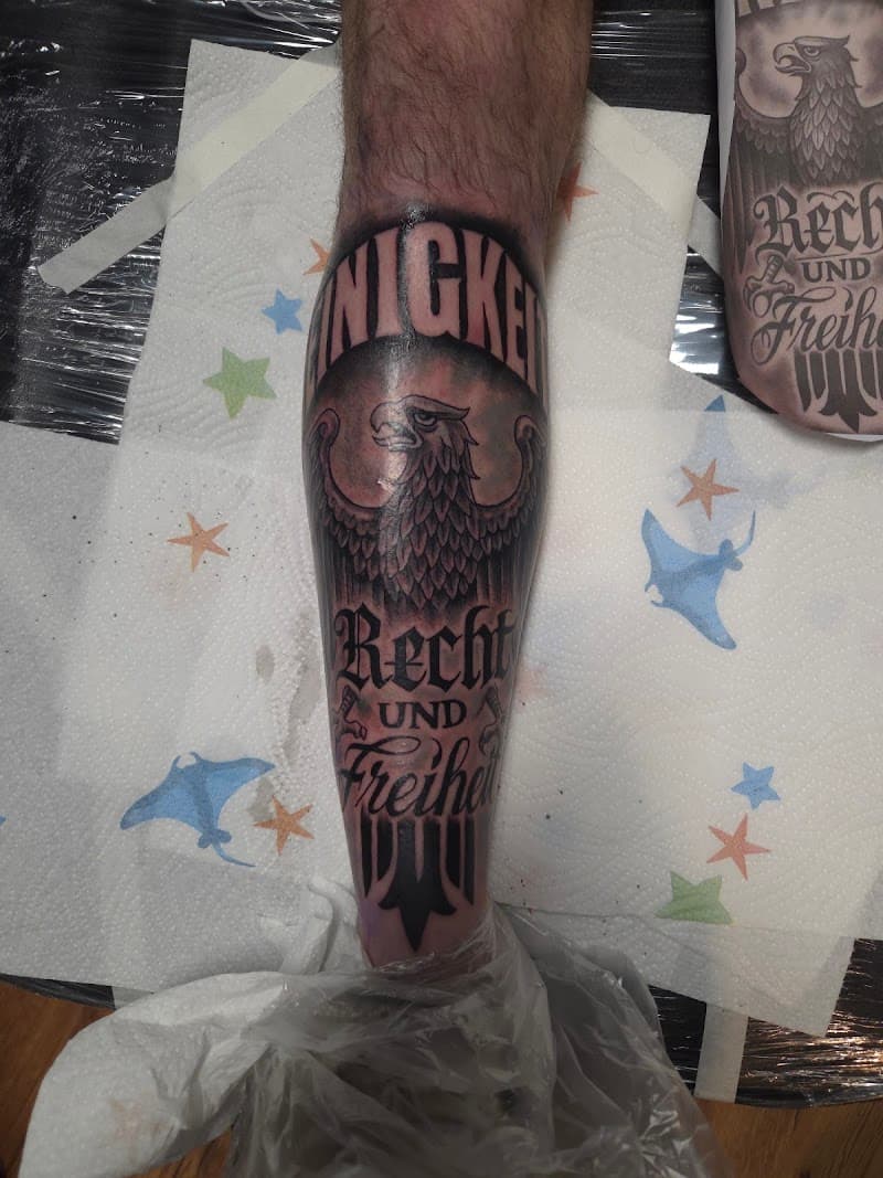 Zwart-grijze adelaar met tekst 'EINIGKEIT RECHT UND FREIHEIT' op onderbeen.