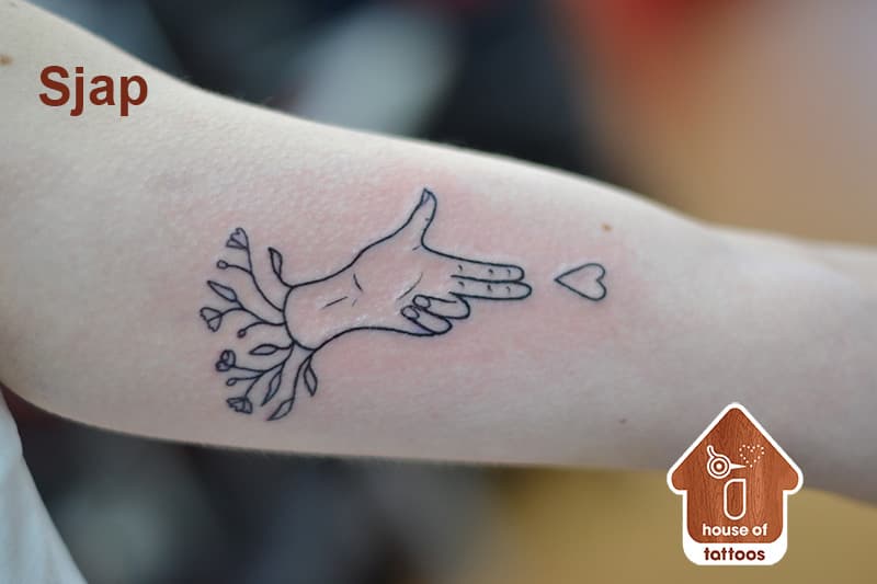 Fineline tattoo van een hand die een hartje 'schiet', met bloemen uit de pols.