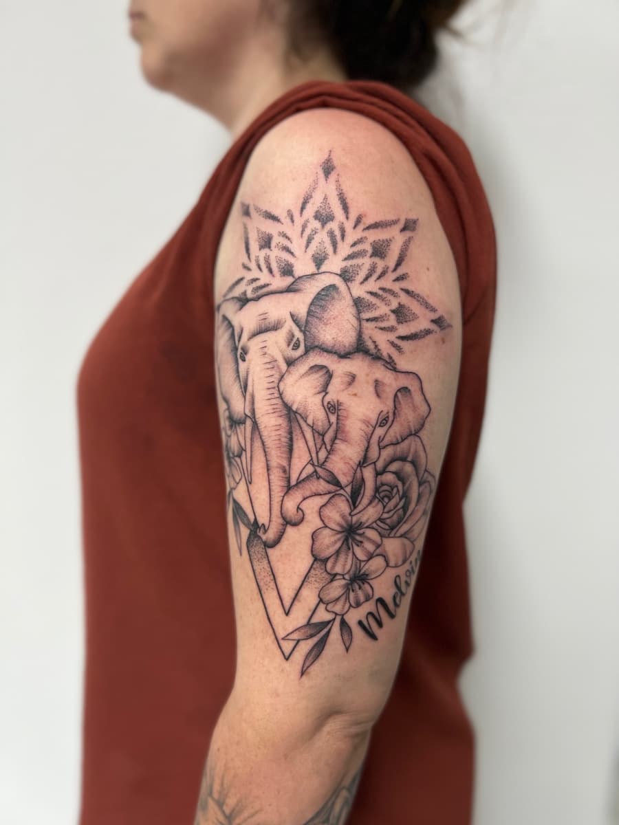 Tattoo van twee olifanten met mandala, bloemen en naam 'Melvin' op bovenarm.