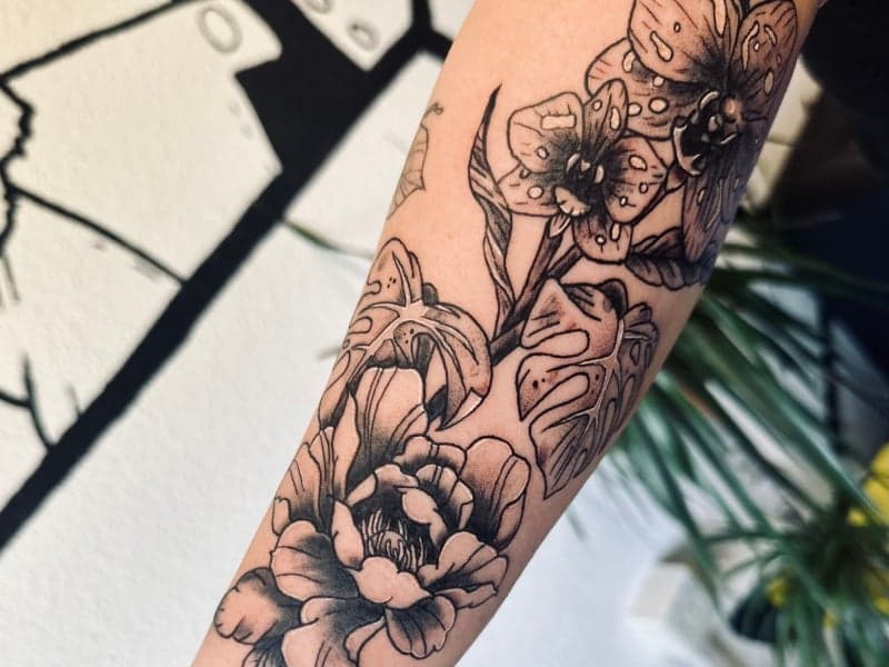 Zwart-wit bloemen tattoo met orchideeën, pioenroos en monstera blad op onderarm.
