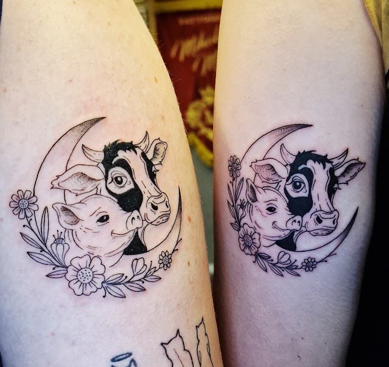 Twee identieke tattoos van een koe en varken in een halve maan met bloemen.