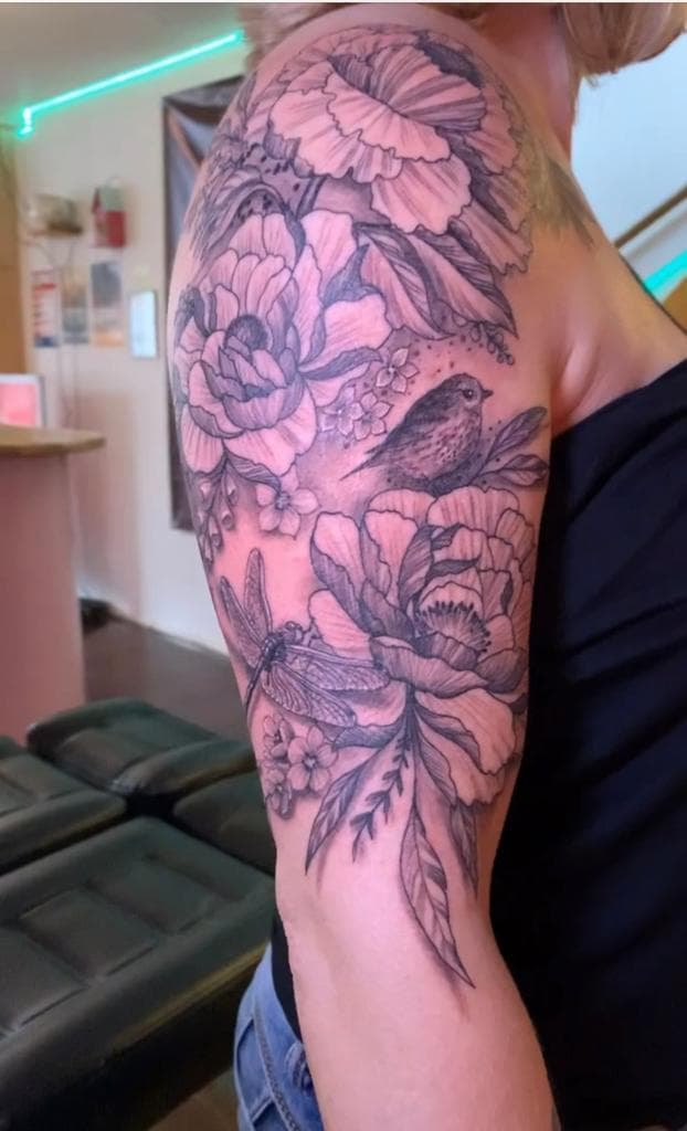 Zwart-wit tattoo van pioenrozen, een vogel en een libel op de bovenarm.