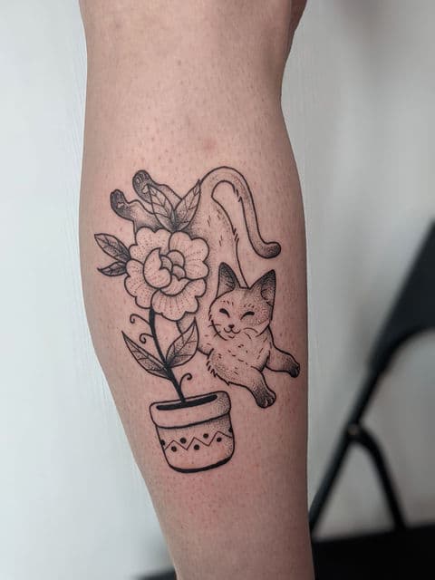 Blackwork tattoo van een speelse kat met een bloem in een pot.