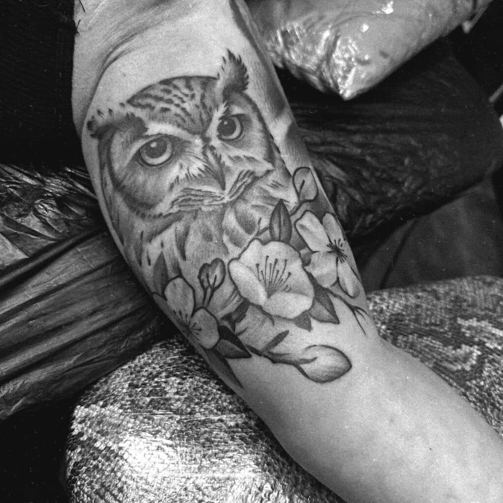 Zwart-wit tattoo van een uil met kersenbloesem op de bovenarm.