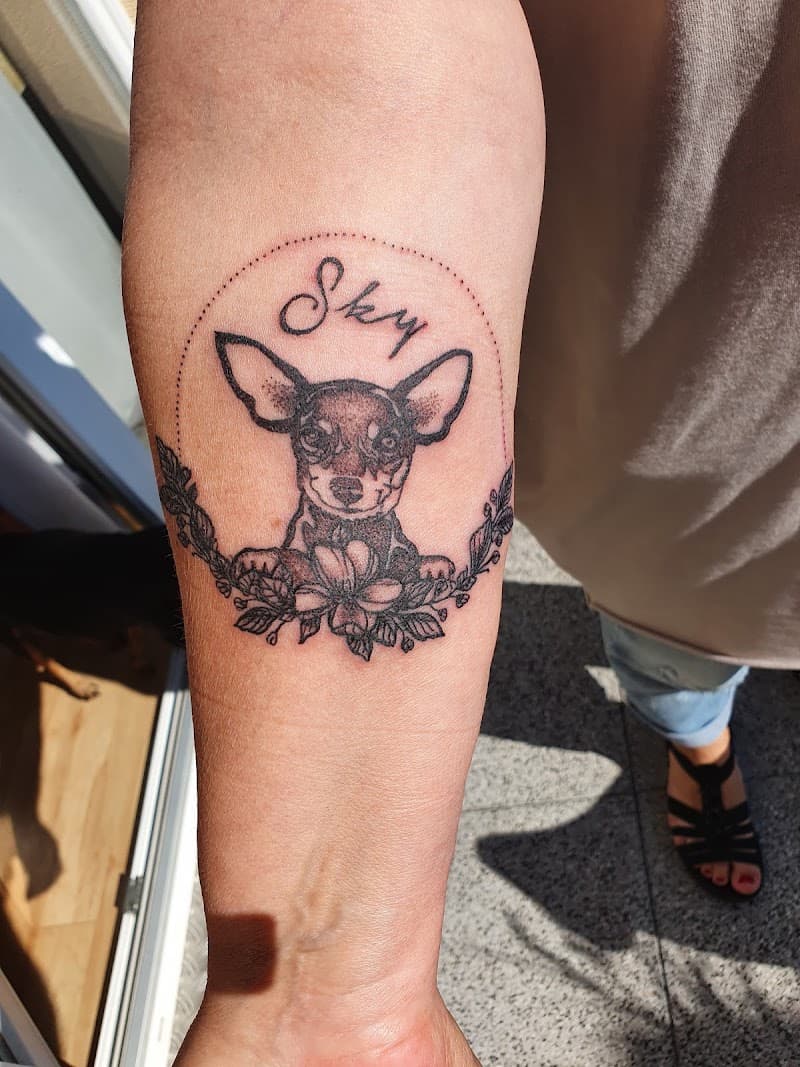 Fineline tattoo van chihuahua 'Sky' met bloemen in cirkel op onderarm.